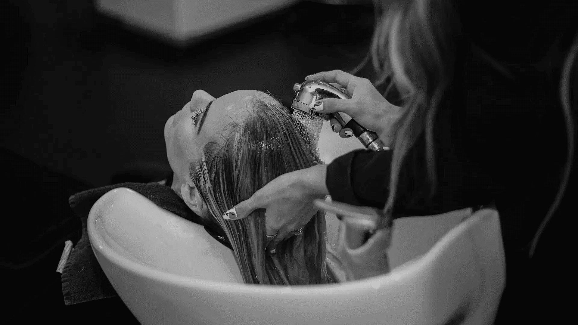Woman getting her hair washed in a salon, laying in a white sink, attended by a hairstylist.